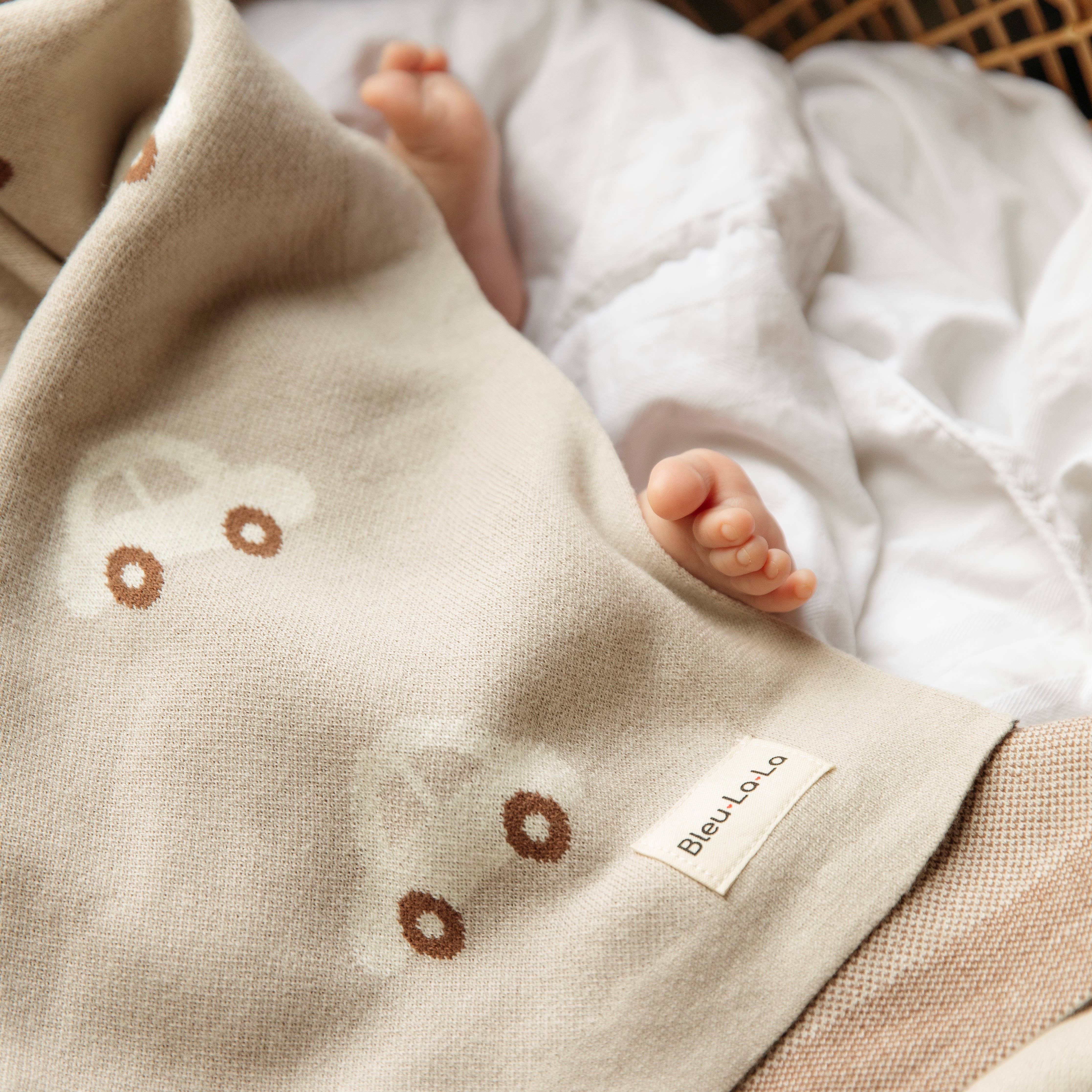 Baby's feet peeking out from under a beige blanket with floral patterns, featuring a 'Bleu Lolo' label.